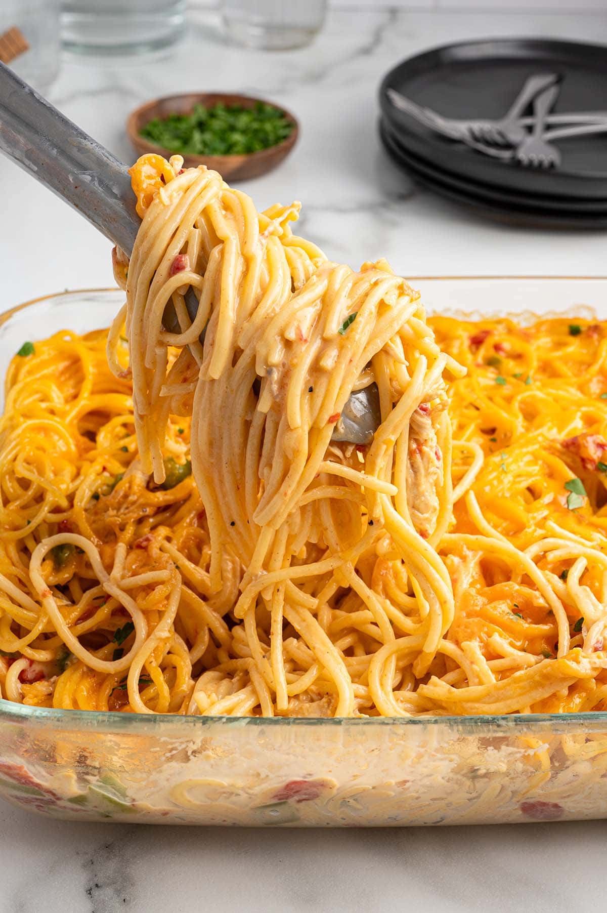 Tongs lifting a serving of Rotel chicken spaghetti from a glass baking dish.