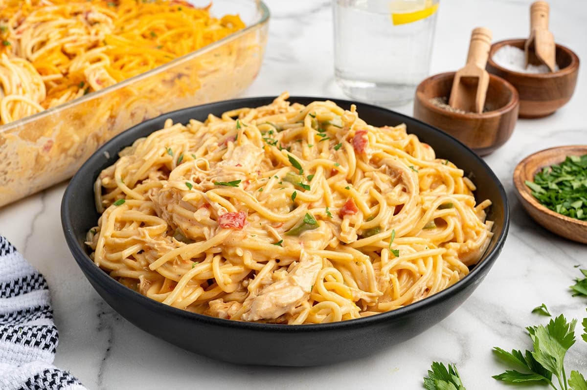 Black bowl of creamy Rotel chicken spaghetti topped with chopped parsley, with the casserole dish in the background.