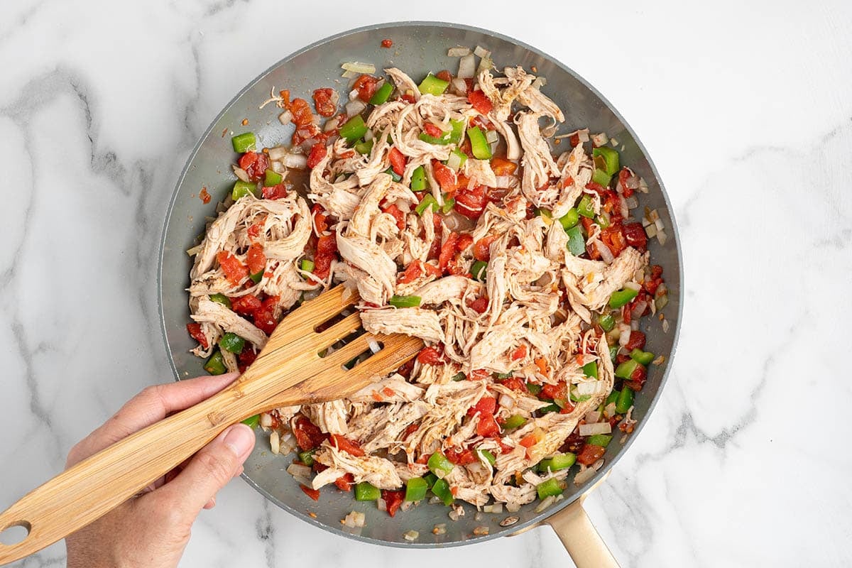 Stirring shredded chicken with diced tomatoes and peppers in a skillet.