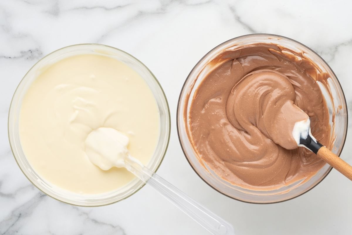 Overhead view of vanilla and chocolate cheesecake batters in separate bowls with mixing spatulas.