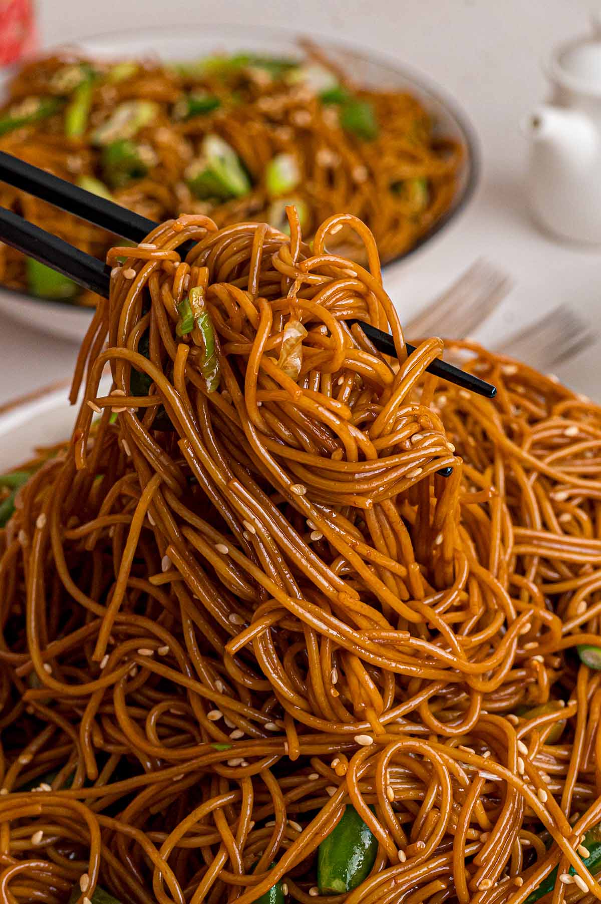 Close-up of sesame-soy dressed Asian Noodle Salad being lifted with black chopsticks, with sesame seeds and green onions throughout.