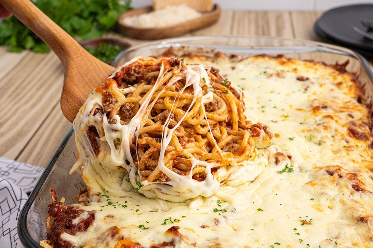 Cheesy baked spaghetti in a casserole dish with a serving lifted on a wooden spoon.