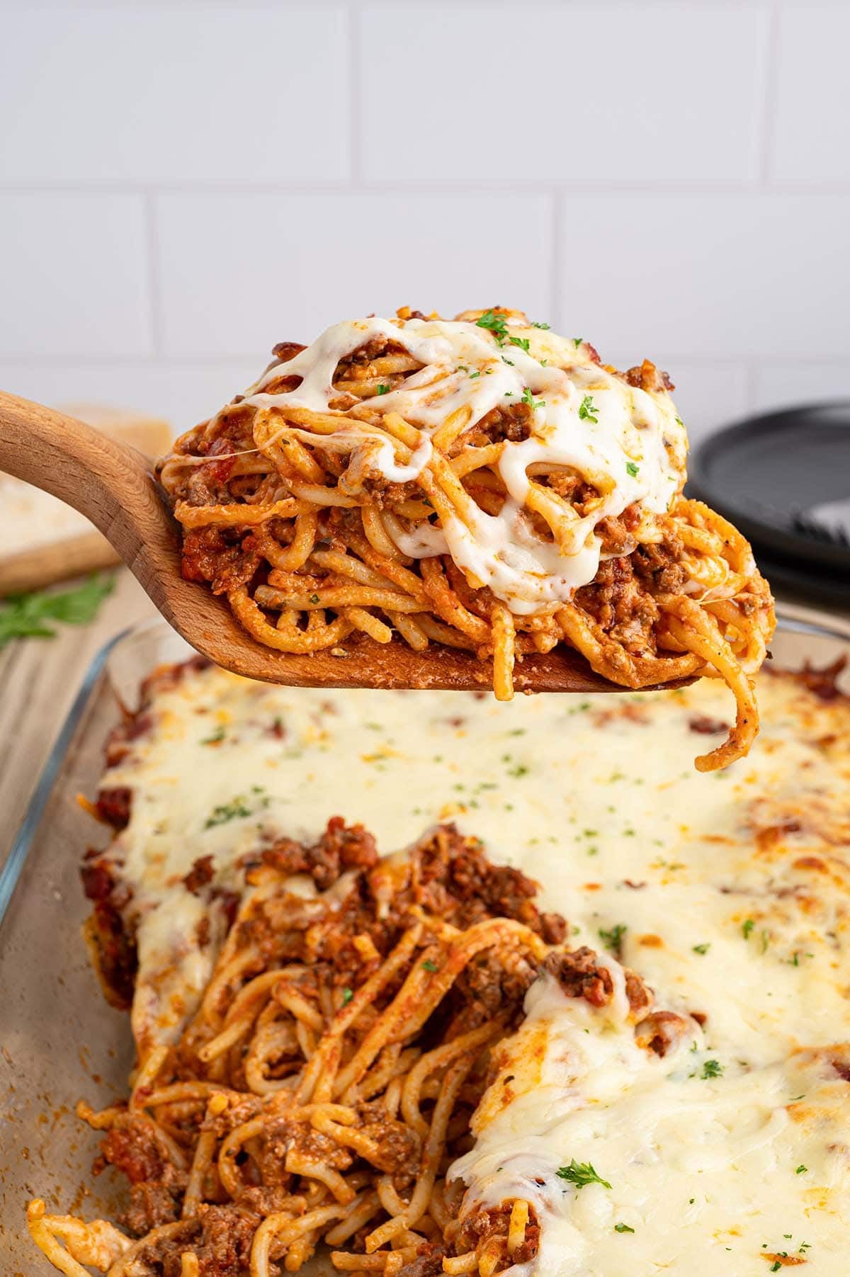 Wooden spoon holding a serving of cheesy baked spaghetti above the casserole dish.