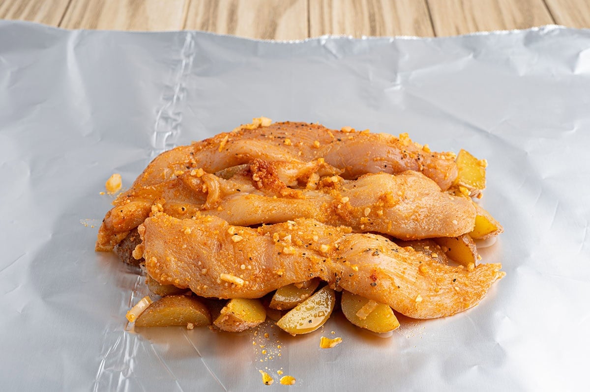 Seasoned chicken, potatoes, and onions piled on a sheet of foil, ready to wrap and cook.