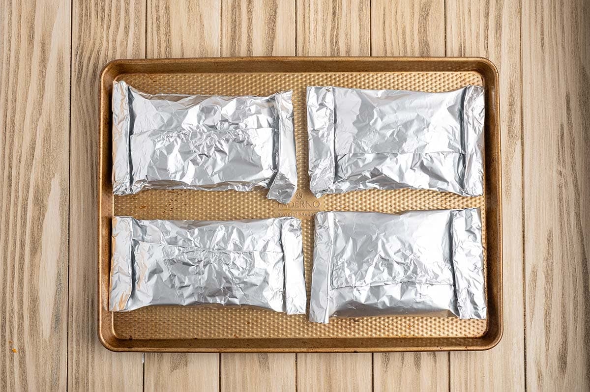 Four sealed chicken foil packets arranged on a baking sheet, ready to bake or grill.
