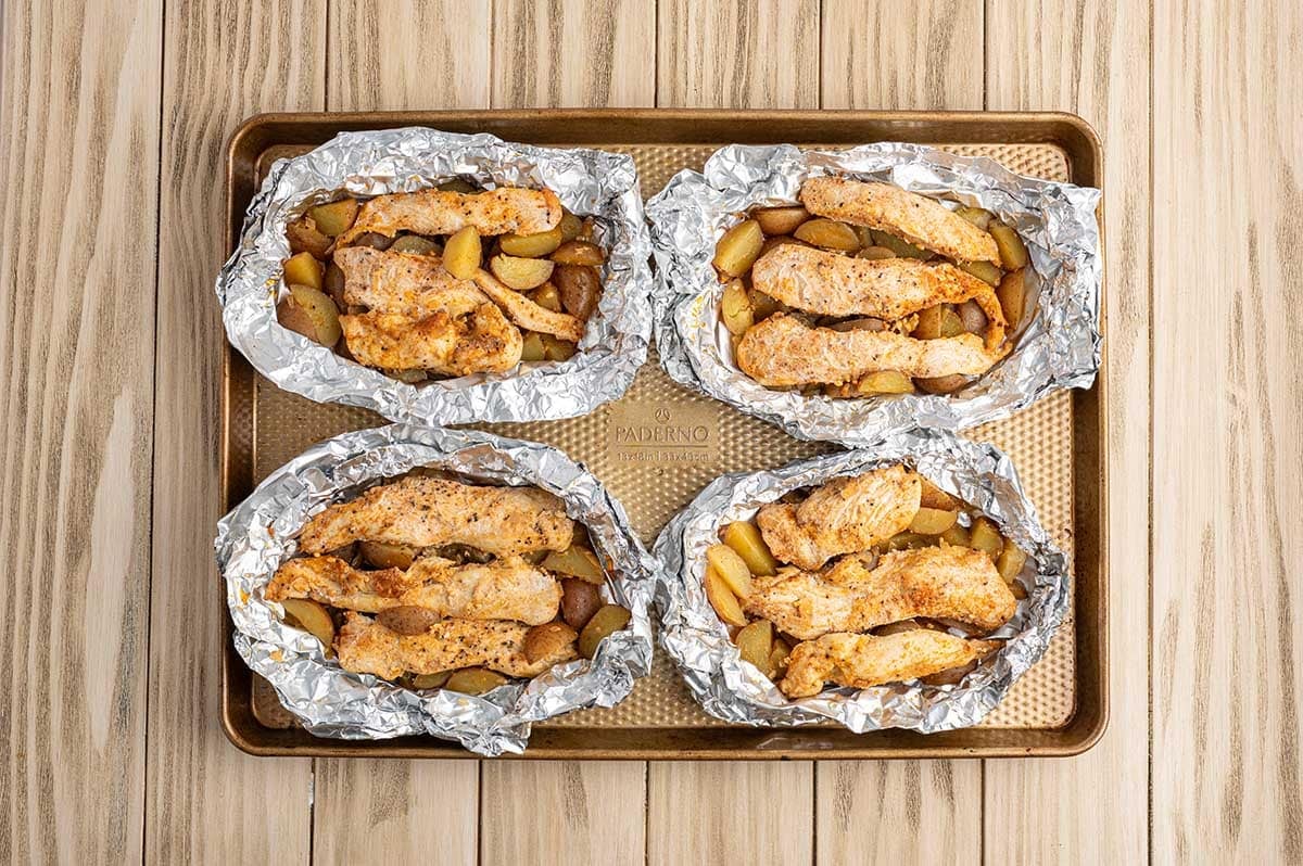 Four open chicken foil packets with cooked chicken and potatoes on a baking sheet, ready for toppings.