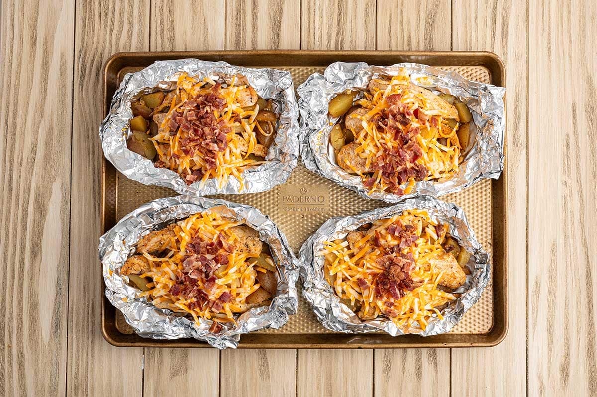 Four open chicken foil packets topped with shredded cheese and crispy bacon on a baking sheet, ready to melt.
