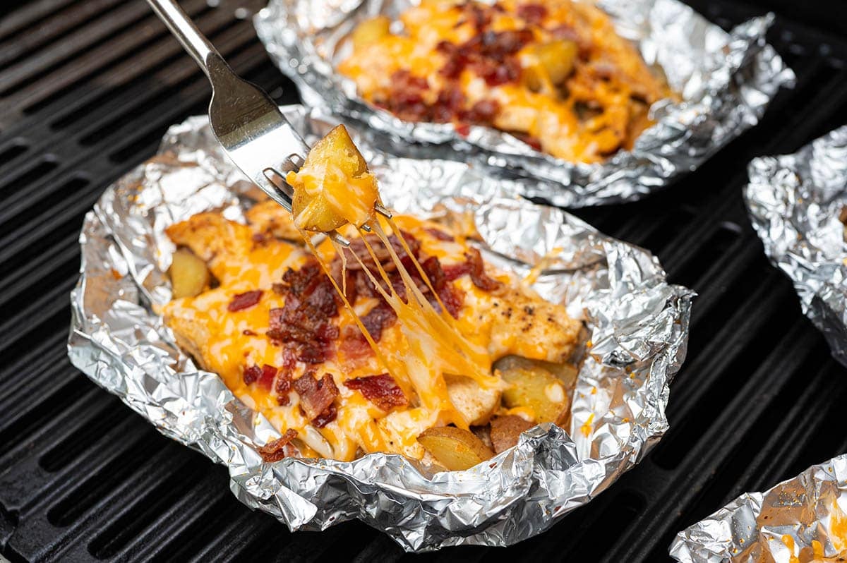 Cheesy bacon chicken foil packet on the grill with a fork lifting a cheesy potato bite.