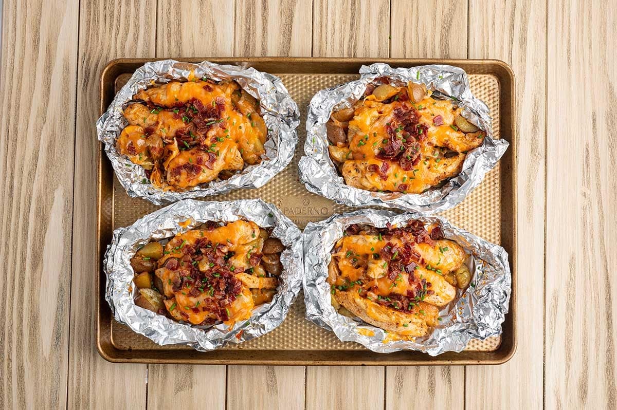 Four cheesy bacon chicken foil packets on a baking sheet, finished with fresh chives and ready to serve.