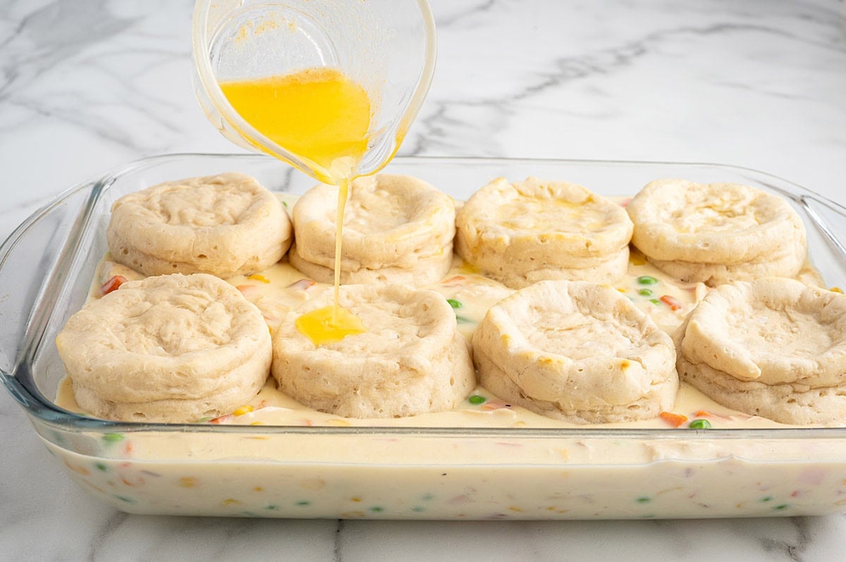 Melted butter being poured over unbaked biscuits on chicken pot pie casserole.