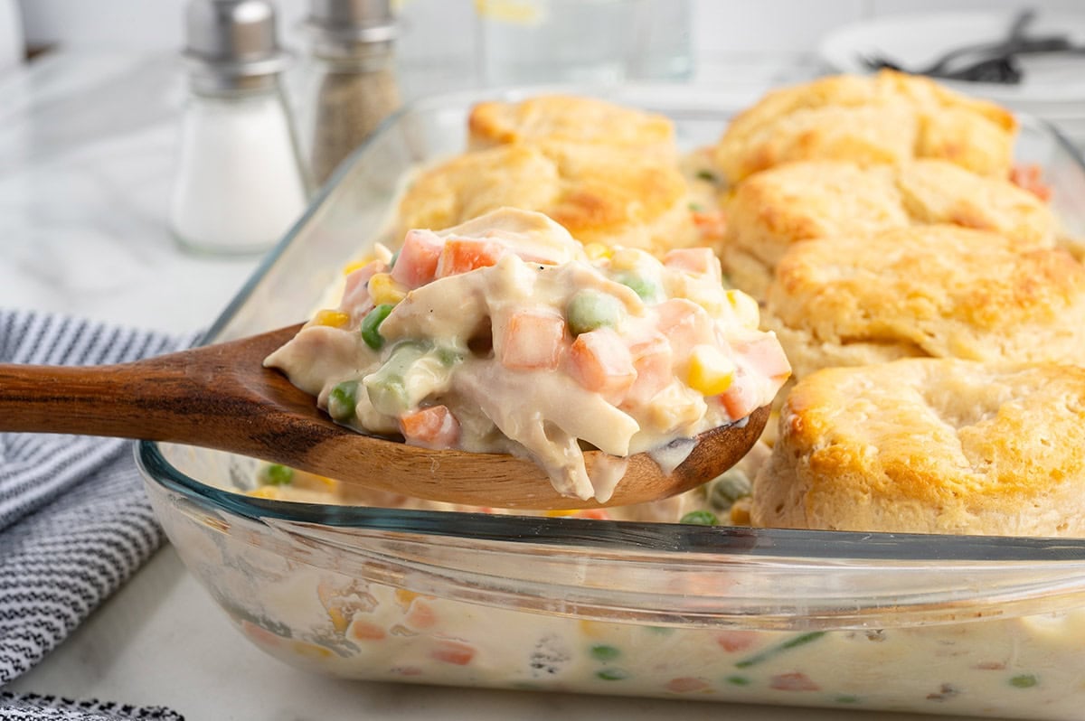 Wooden spoon scooping creamy chicken pot pie casserole with a biscuit topping.