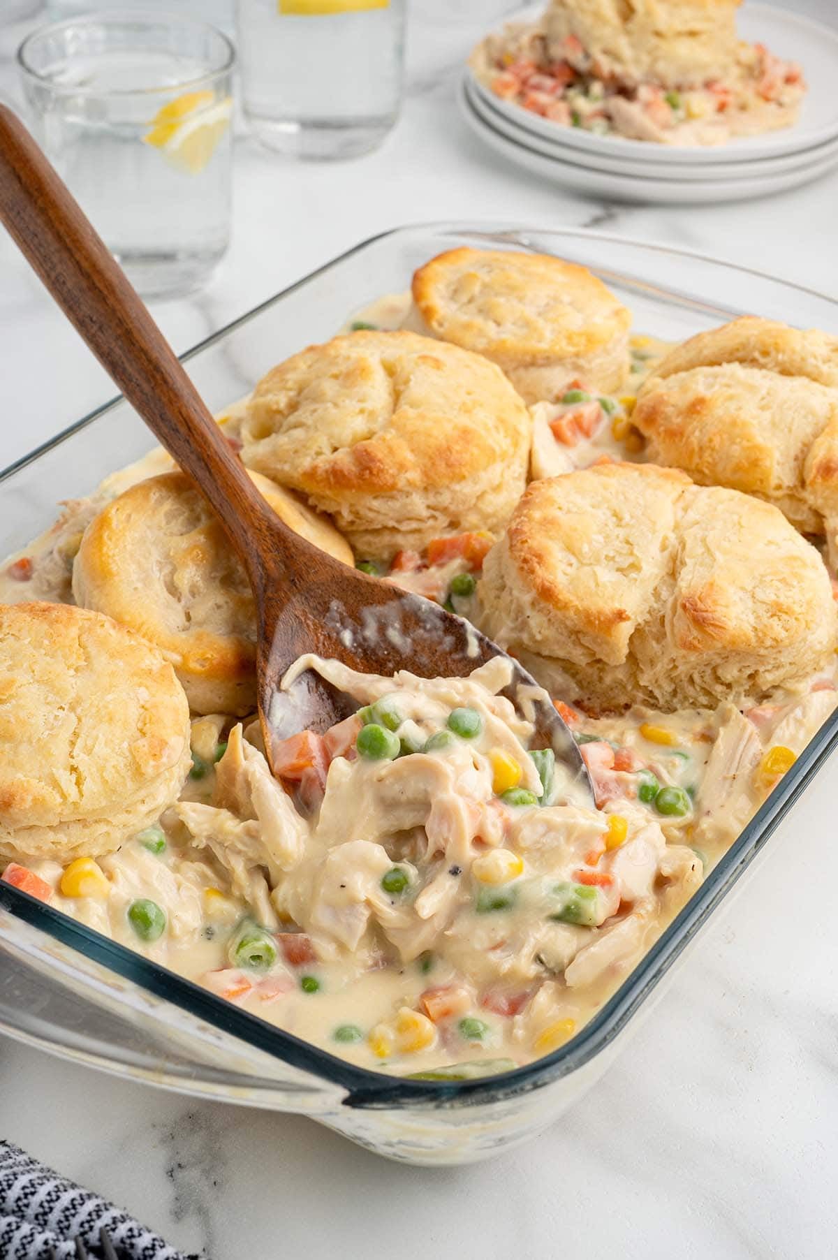 Wooden spoon lifting shredded chicken and vegetables from a biscuit-topped pot pie casserole.