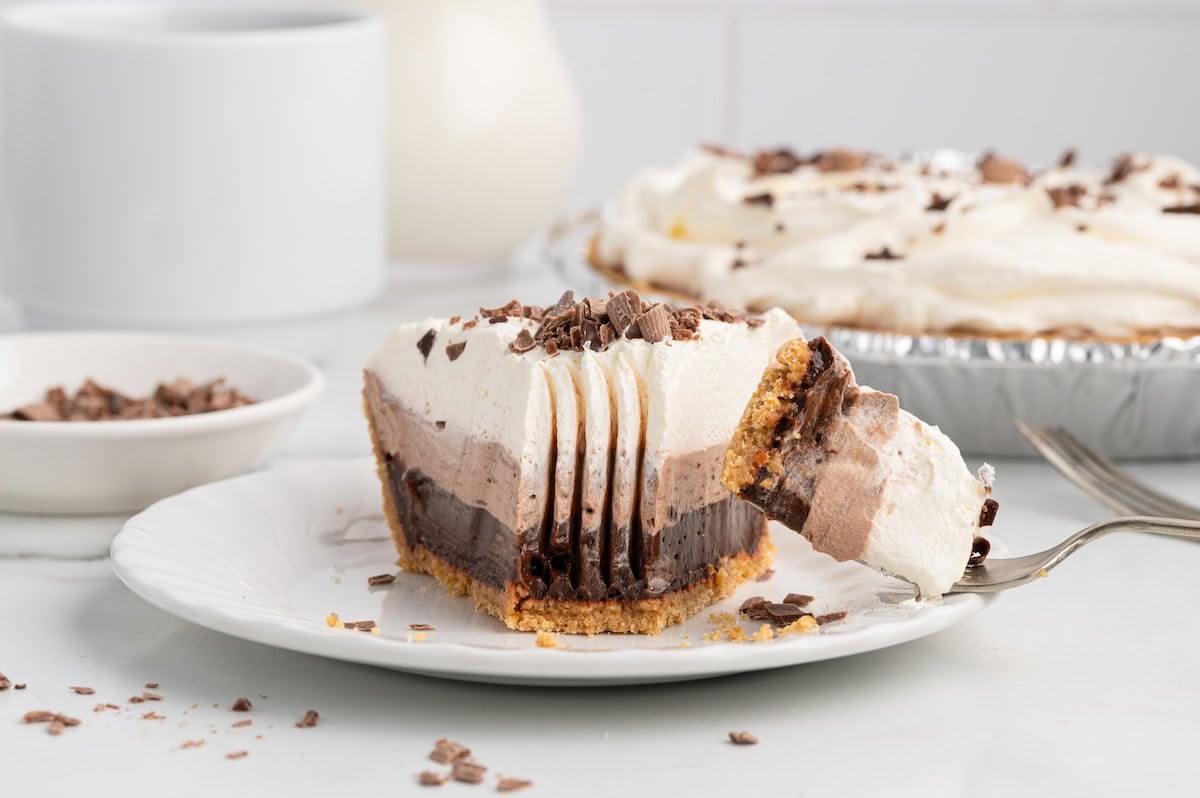 Fork lifting a bite from a slice of layered chocolate pudding pie with whipped cream.