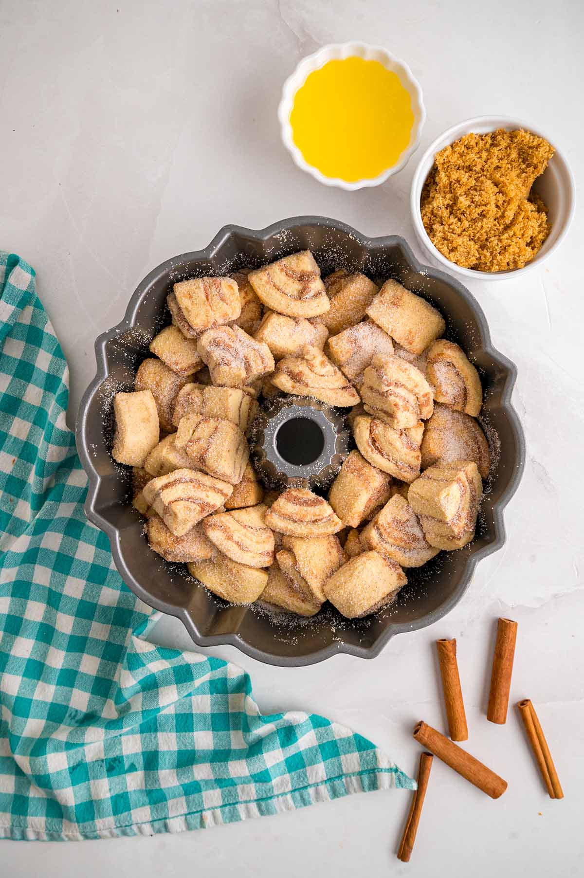 Cinnamon sugar coated cinnamon roll pieces arranged in a Bundt pan with melted butter and brown sugar nearby.