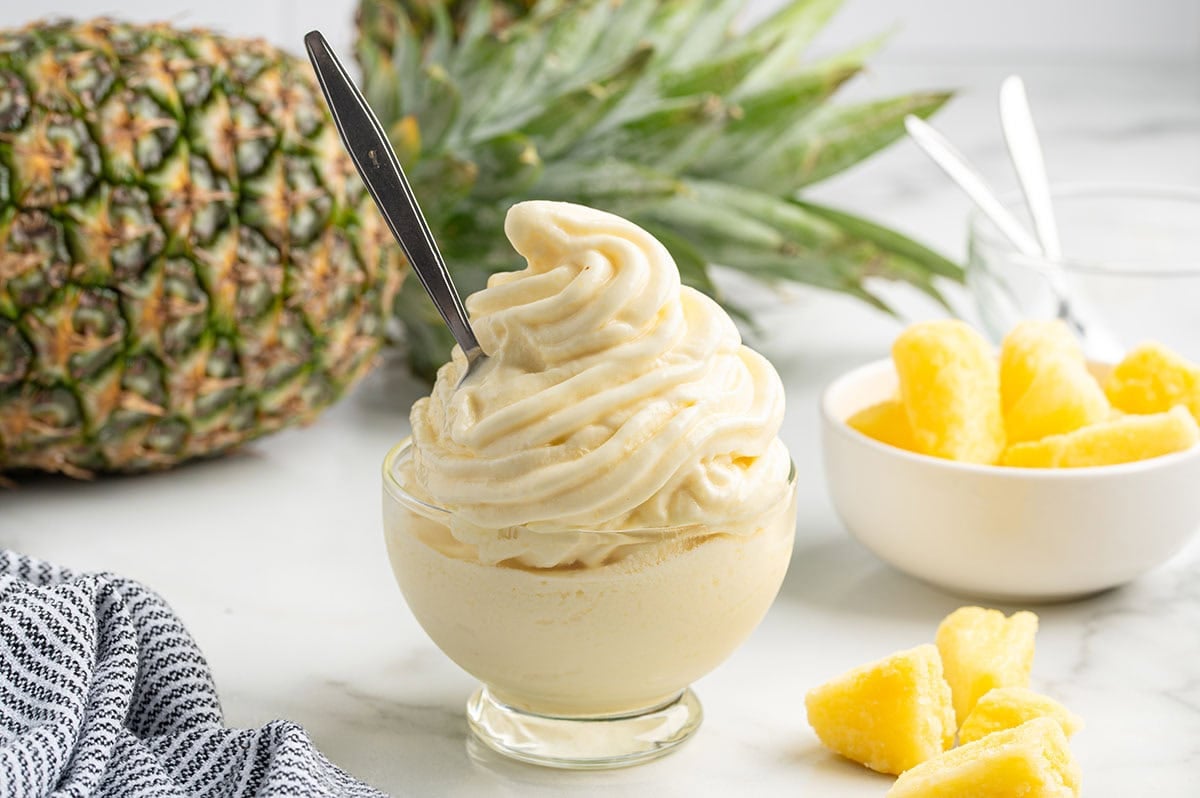 Pineapple Dole Whip swirled in a glass dish with a spoon, next to a whole pineapple and bowl of pineapple chunks