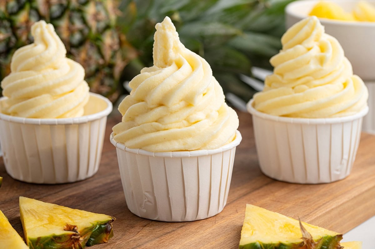 Three cups of Dole Whip pineapple soft serve swirled high, surrounded by fresh pineapple wedges on a wooden board.