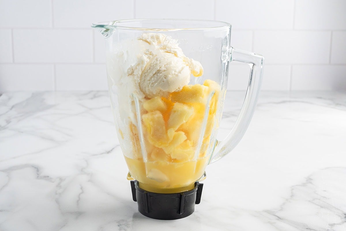 Blender pitcher filled with vanilla ice cream, frozen pineapple chunks, and pineapple juice, ready to blend.