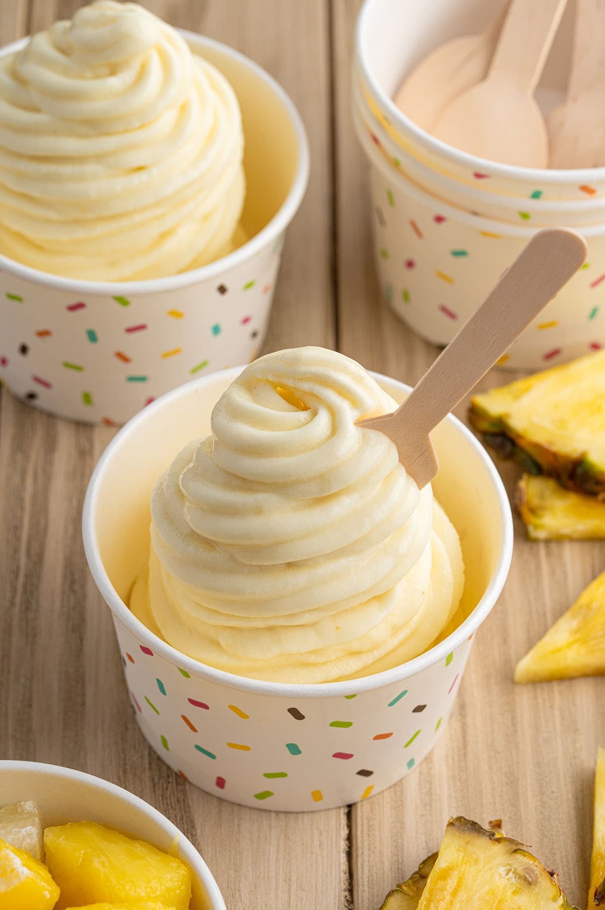 Pineapple Dole Whip swirled in a confetti-print cup with a wooden spoon, surrounded by fresh pineapple chunks.