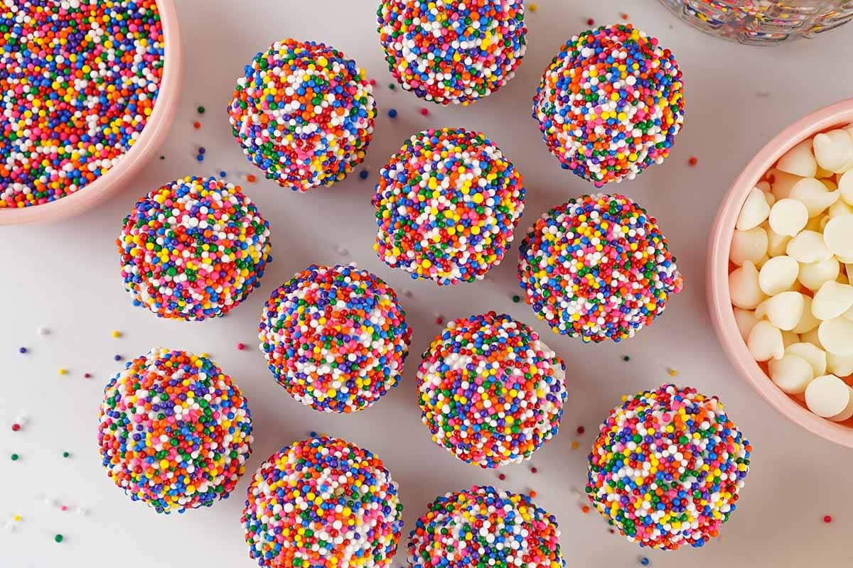 Colorful sprinkle coated cake bites arranged on a surface with bowls of sprinkles and white chocolate chips.