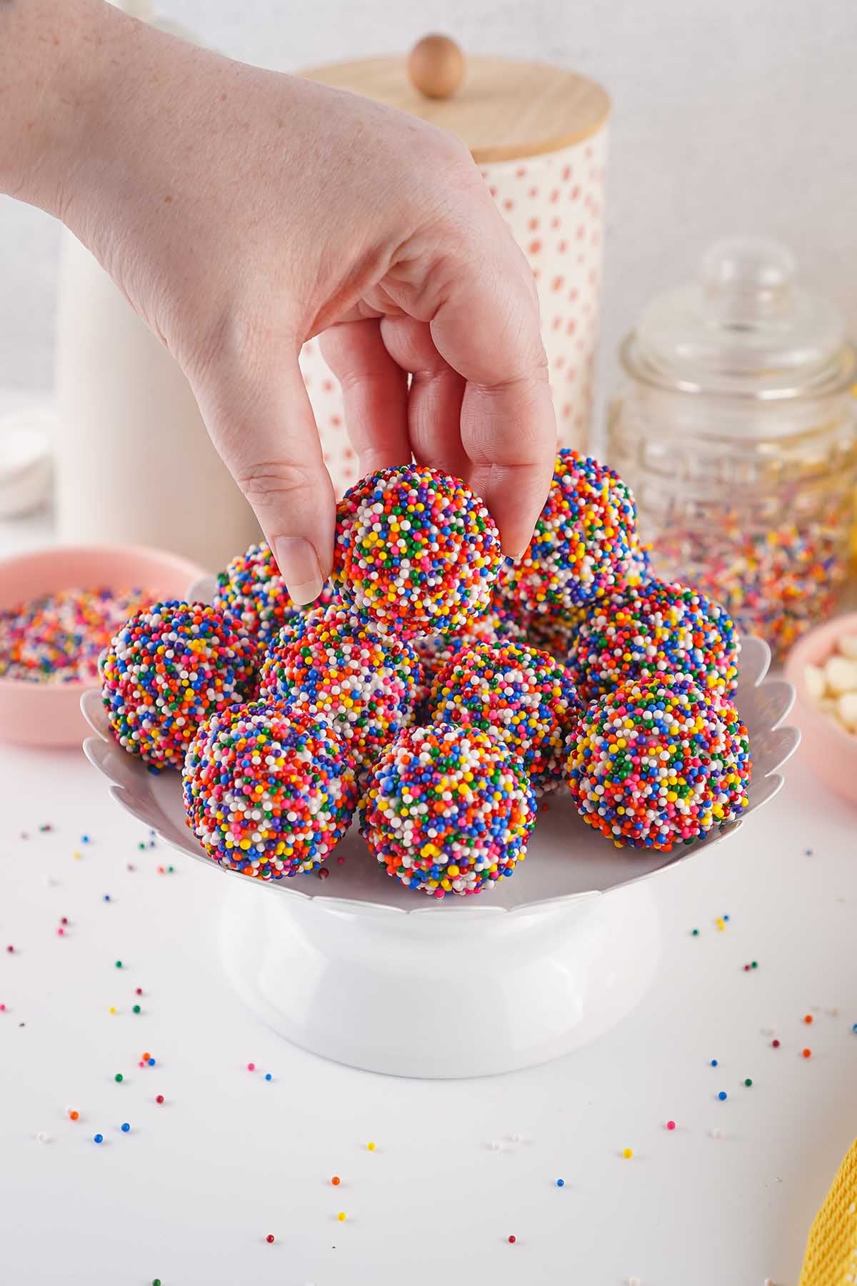 Hand picking up a colorful sprinkle coated cake bite from a serving plate.