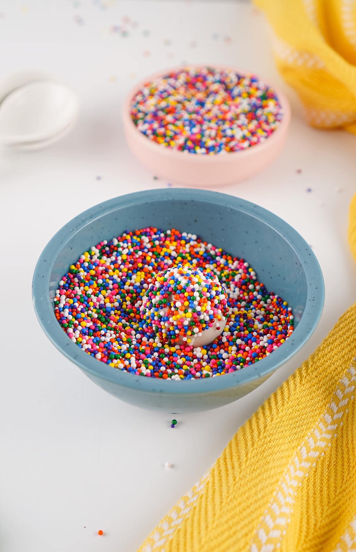 Cake ball coated with rainbow sprinkles in a bowl of nonpareils.