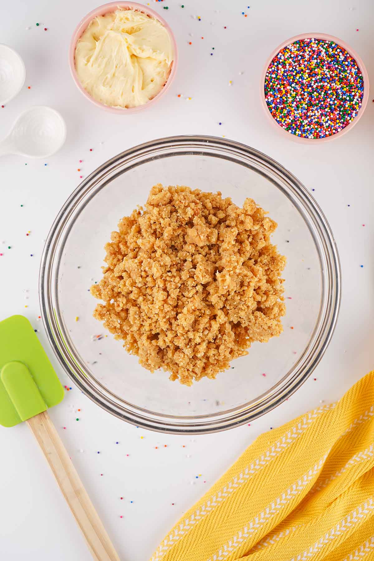 Crumbled cake mixture in a glass mixing bowl with frosting, rainbow nonpareils, and a spatula ready to make cake bites.
