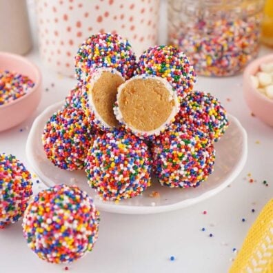Dot Cake Bites covered in rainbow nonpareil sprinkles on a white plate, with one cut in half showing the dense vanilla cake interior.