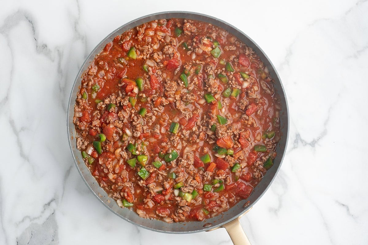 Ground beef simmering with diced tomatoes and green peppers in a skillet.