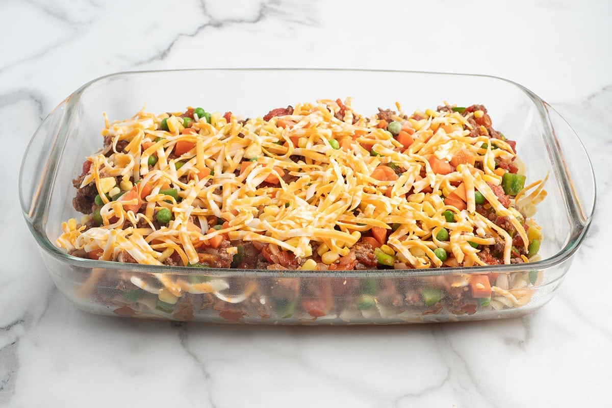 Hamburger casserole topped with shredded cheese in a baking dish before baking.