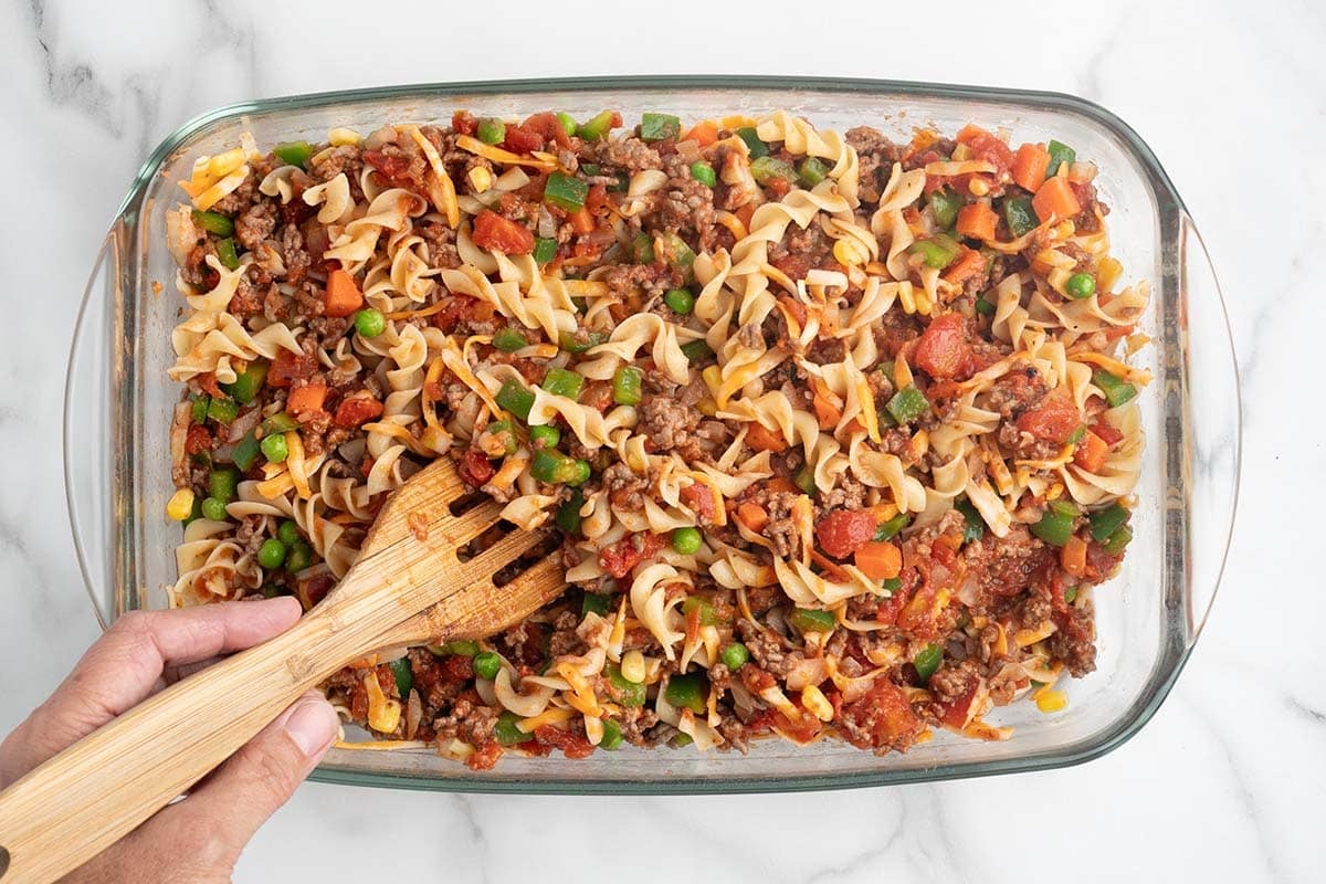 Hand mixing hamburger casserole with egg noodles and vegetables with a wooden spoon.