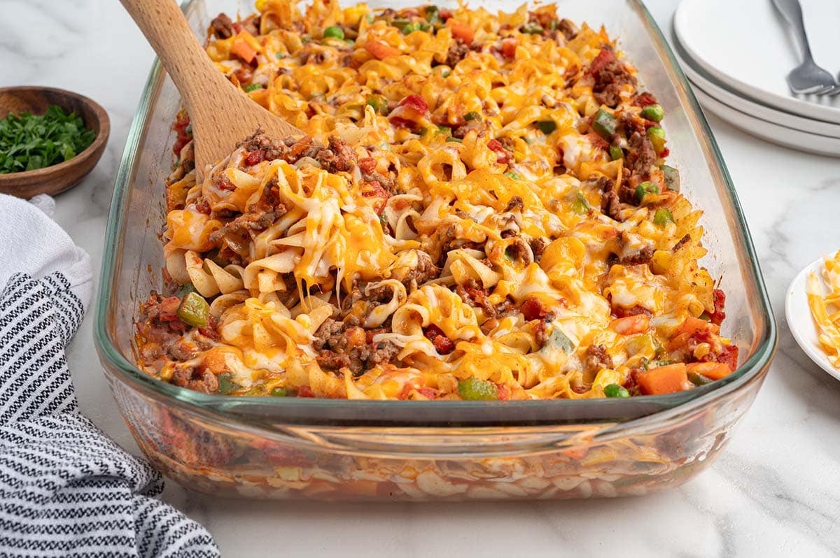 Wooden spoon lifting cheesy hamburger casserole with egg noodles and vegetables from the dish.