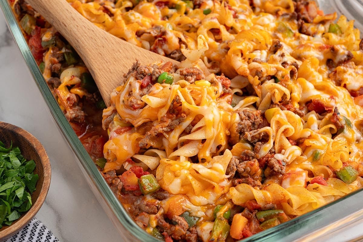 Wooden spoon scooping cheesy hamburger casserole with egg noodles and vegetables.