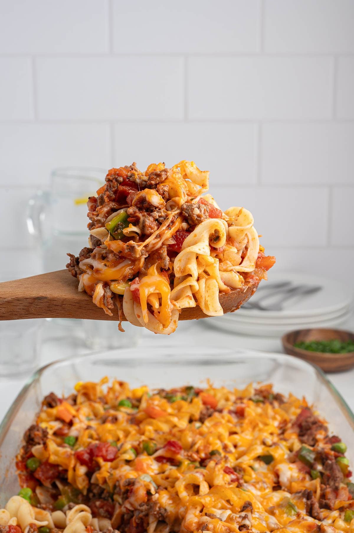 Wooden spoon lifting a scoop of cheesy hamburger casserole with egg noodles above the baking dish.