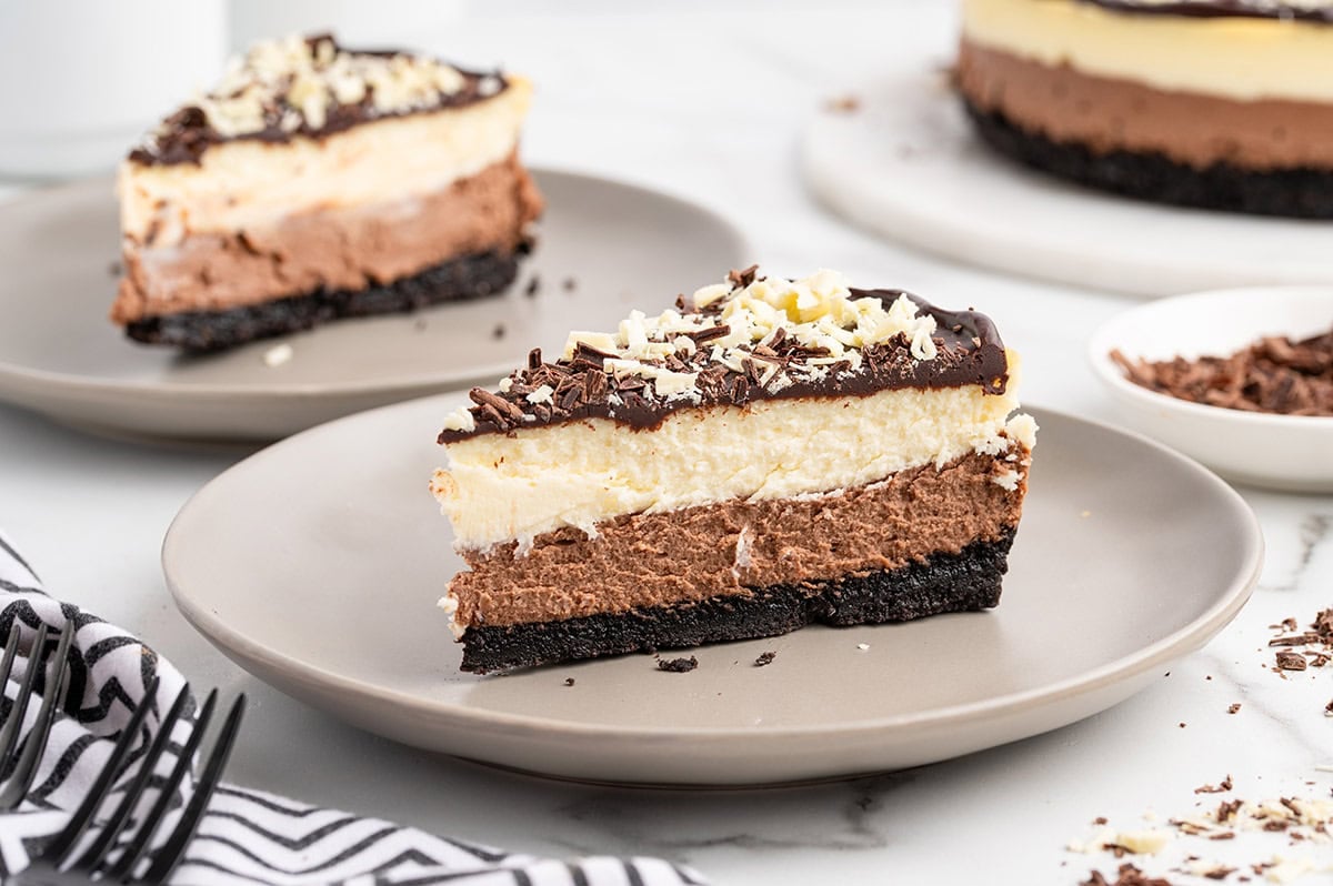 Slice of layered chocolate cheesecake with an Oreo crust, chocolate and vanilla layers, topped with ganache and chocolate shavings.