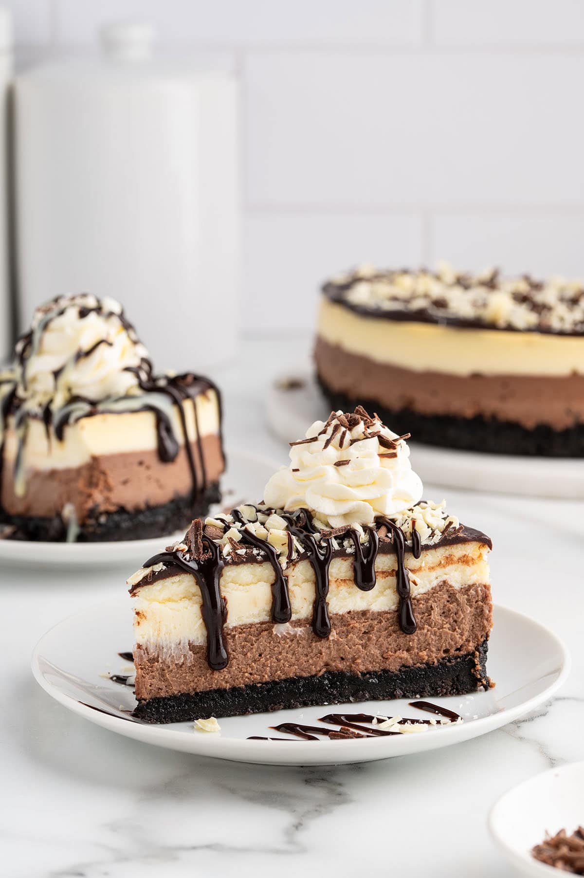 Slice of layered chocolate cheesecake drizzled with chocolate sauce and topped with whipped cream and chocolate shavings.