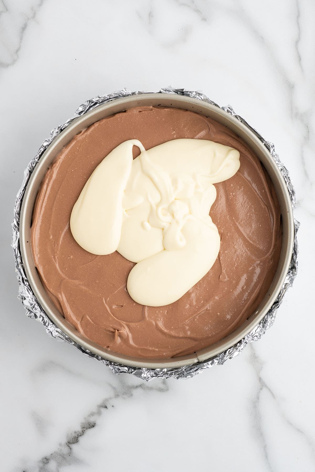 Overhead view of vanilla cheesecake batter poured over chocolate batter in a foil-wrapped springform pan.