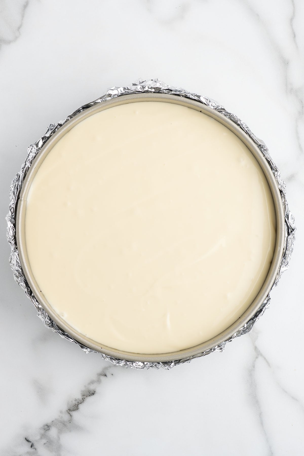 Overhead view of smooth vanilla cheesecake batter spread evenly in a foil-wrapped springform pan.