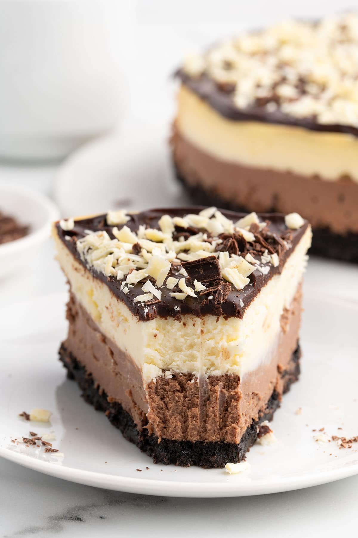 Close-up of a layered chocolate cheesecake slice with Oreo crust, chocolate and vanilla layers, ganache, and chocolate shavings on top