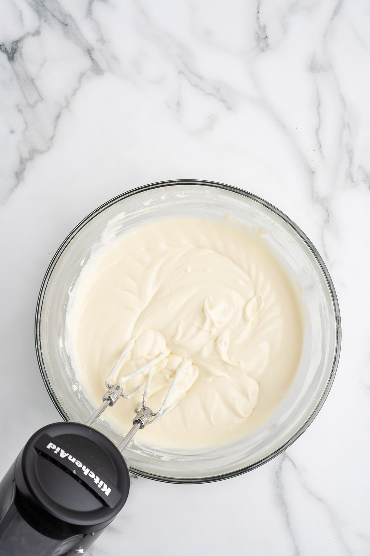 Overhead view of cream cheese batter being mixed in a glass bowl with a hand mixer.