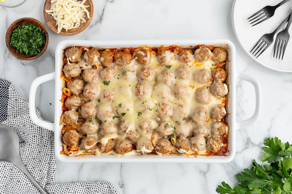 Baked meatball casserole with melted cheese in a white dish surrounded by parsley and forks.