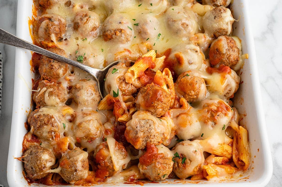 Spoon lifting a cheesy serving of meatball pasta casserole from the baking dish.
