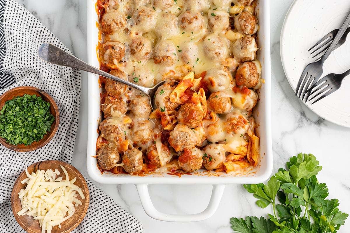 Overhead shot of meatball casserole with a spoon, surrounded by parsley, cheese, and forks.