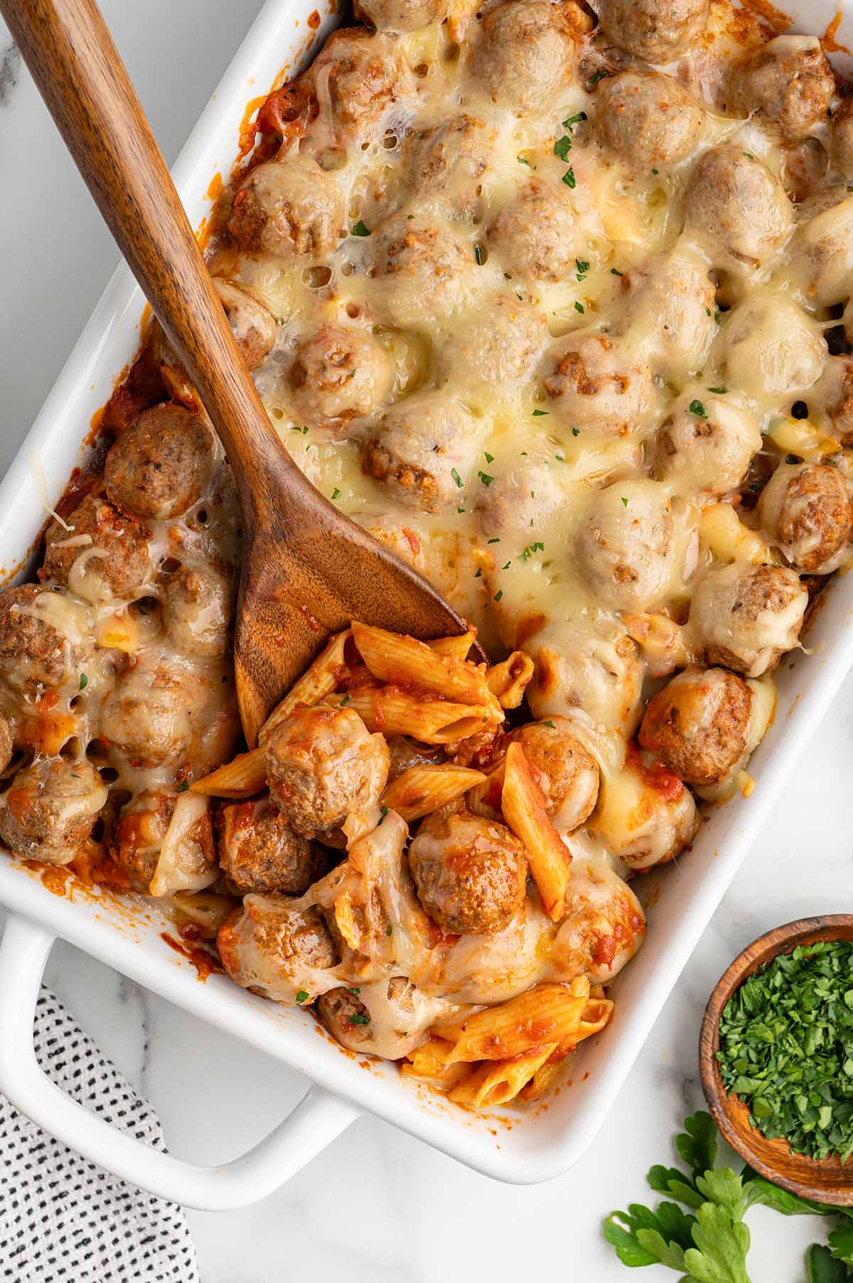 Overhead view of cheesy meatball casserole with a wooden spoon serving up penne pasta.