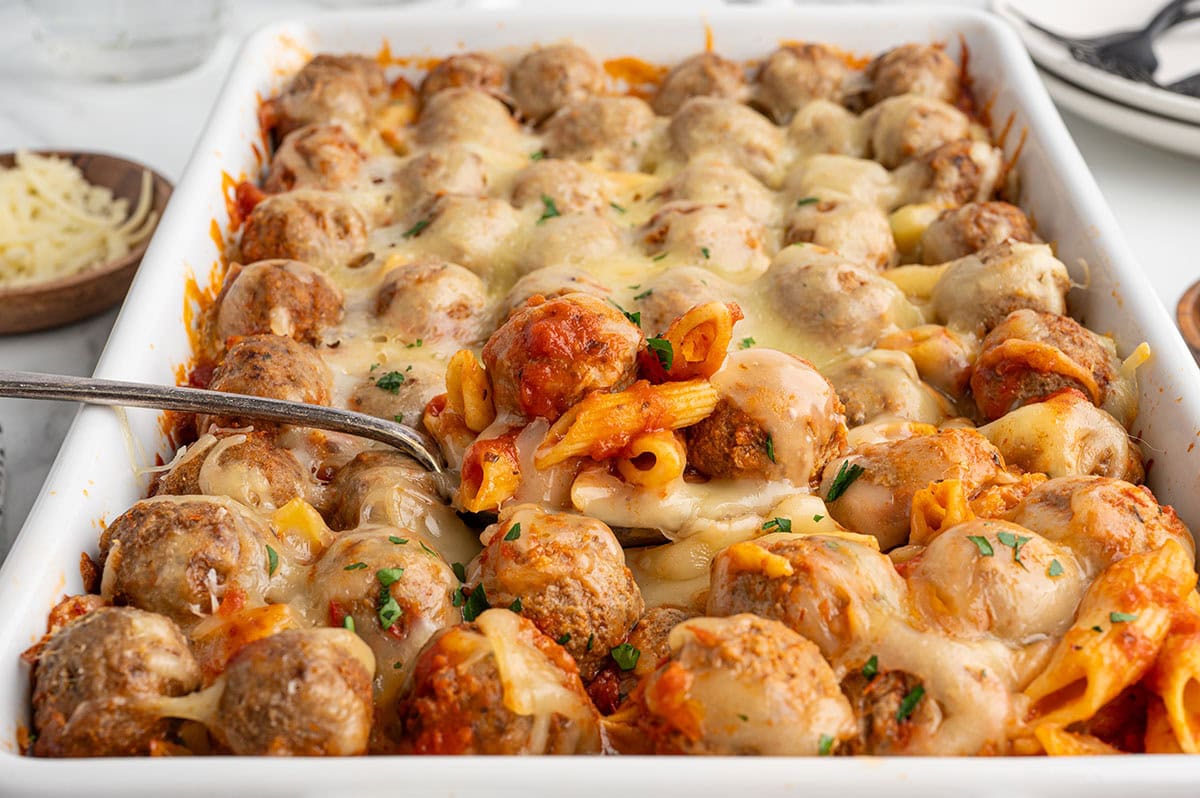 Cheesy meatball pasta casserole in a white baking dish with a spoon lifting a serving.