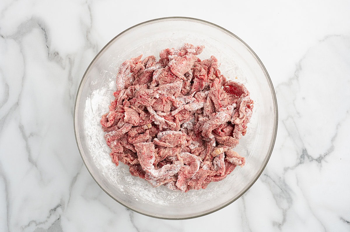 Raw beef strips coated in cornstarch in a glass bowl on a marble surface.