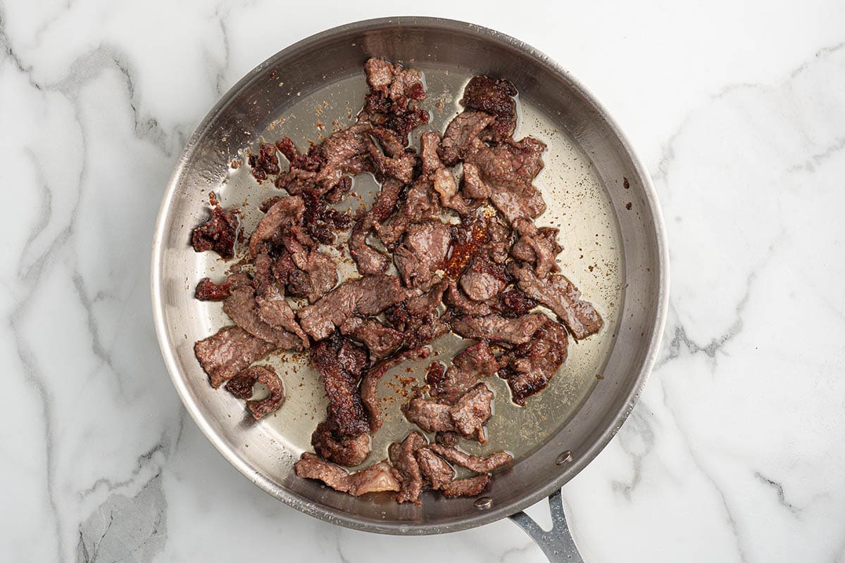 Seared beef strips cooking in a stainless steel skillet on a marble surface.