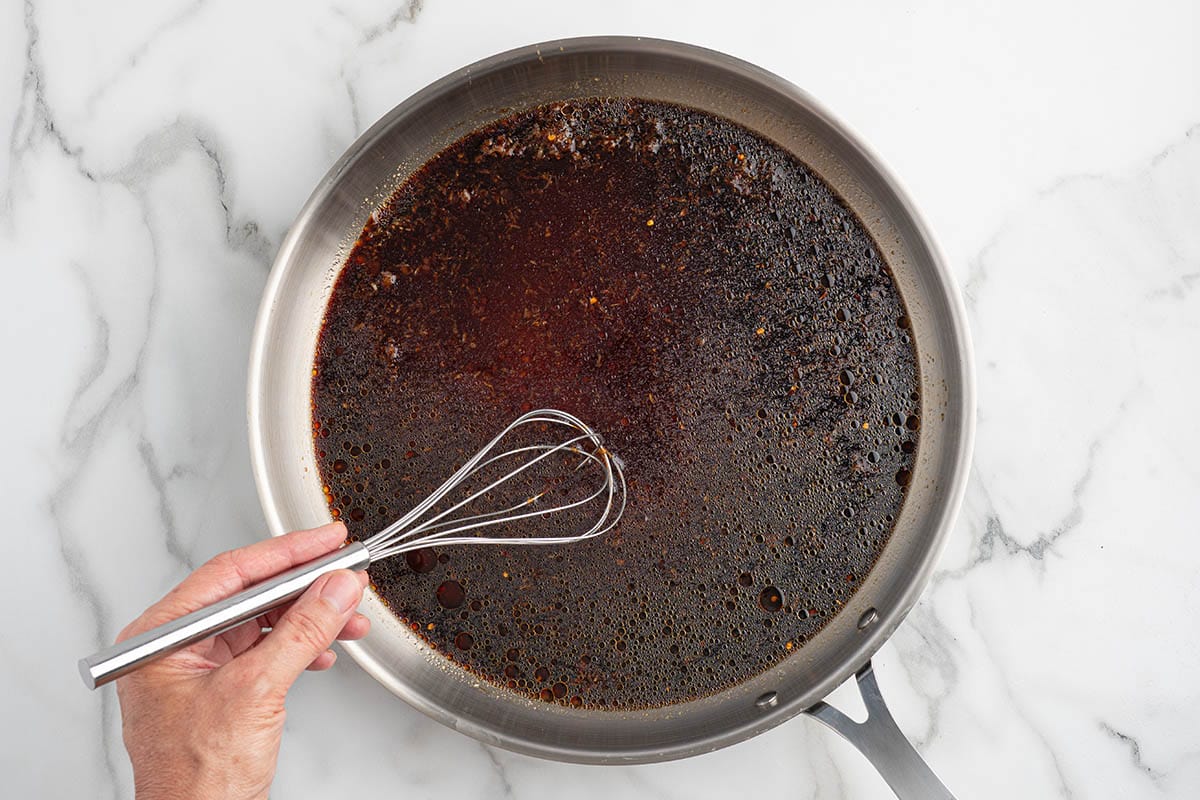 Whisking a dark savory sauce in a stainless steel skillet on a marble surface.