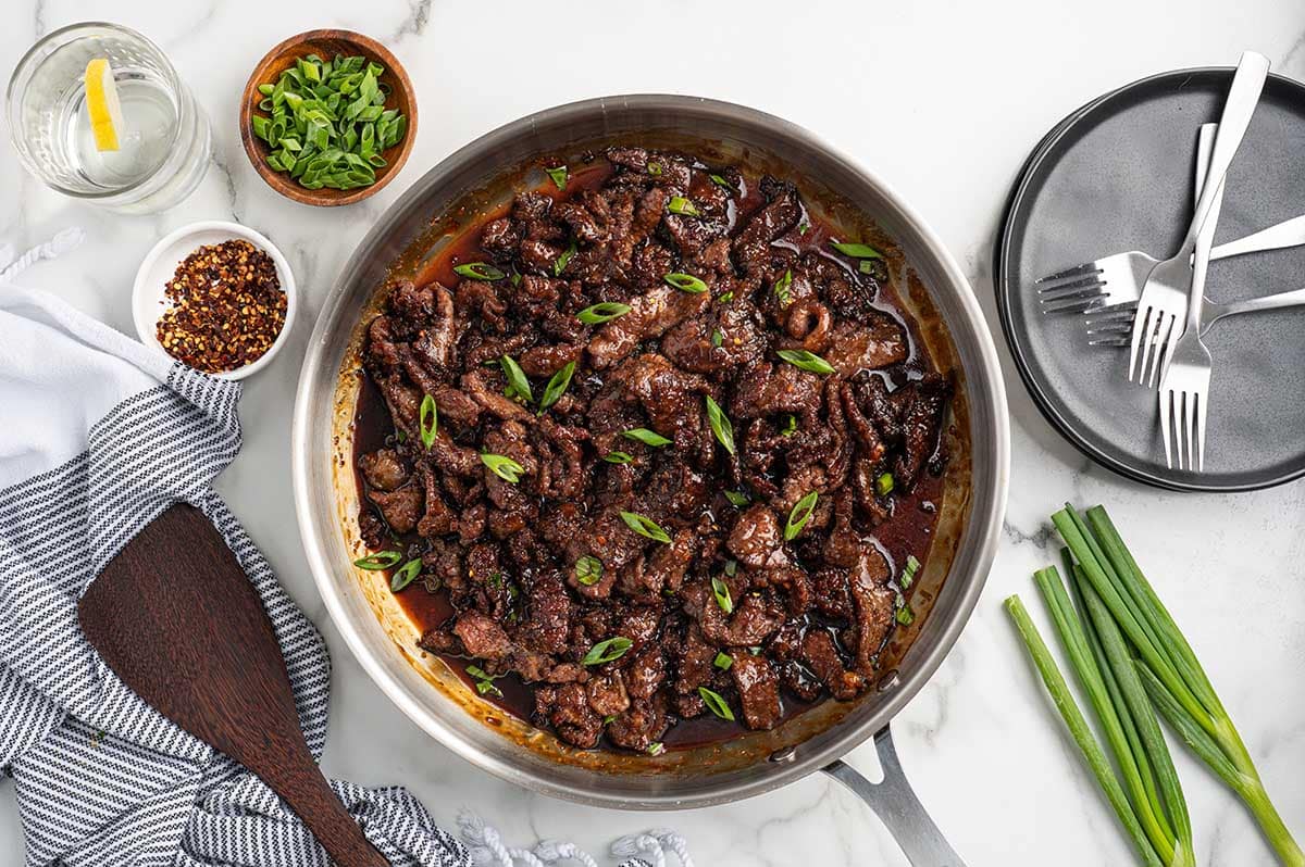 Skillet of Mongolian beef topped with green onions, surrounded by scallions, red pepper flakes, plates, and forks.
