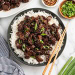 Mongolian beef with green onions served over white rice on a dark plate, with chopsticks, a bowl of chopped scallions, and red pepper flakes nearby.