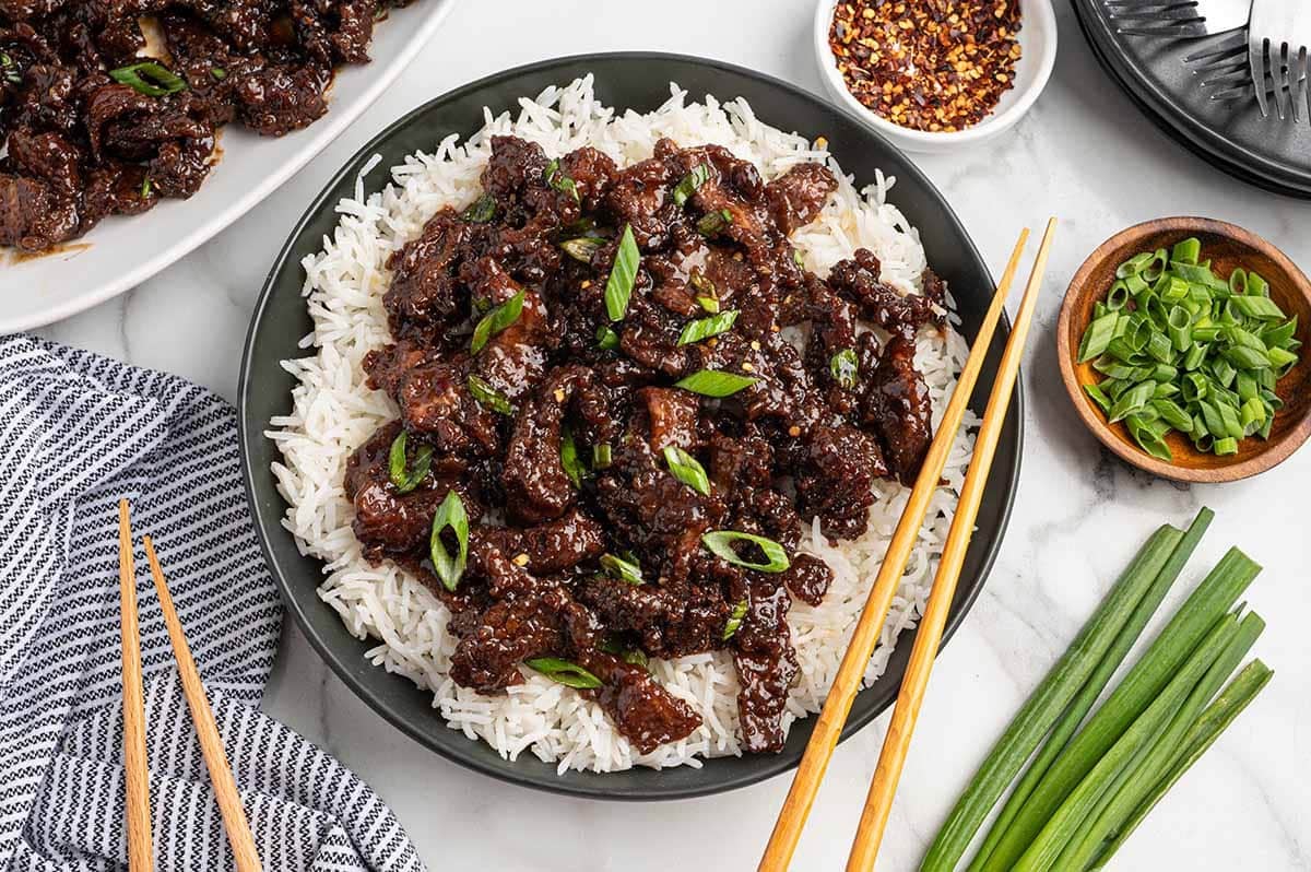 Mongolian beef with green onions served over white rice on a dark plate, with chopsticks, a bowl of chopped scallions, and red pepper flakes nearby.